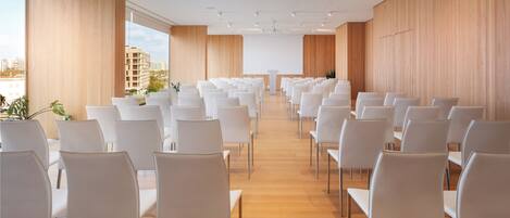 A modern conference room with rows of white chairs, wooden floors, and large windows offering a city view.