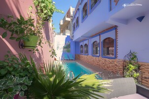 Zona de piscina con un edificio azul y rosa, dos sillones de descanso y plantas en macetas.