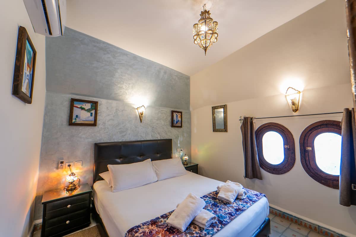 Quarto de hotel com cama, mesas de cabeceira, luminárias na parede, um candelabro e duas janelas com cortinas.