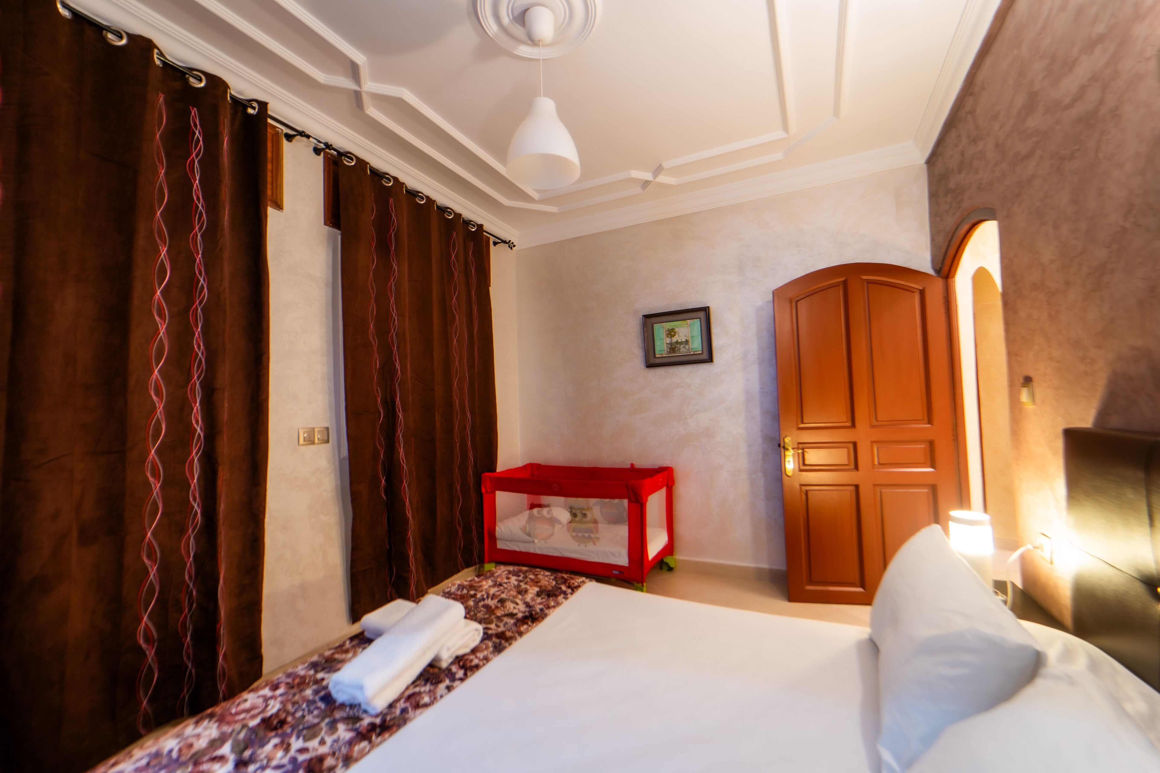 Une chambre d’hôtel avec un lit, une boîte de rangement rouge, une armoire en bois et un tableau encadré au mur.