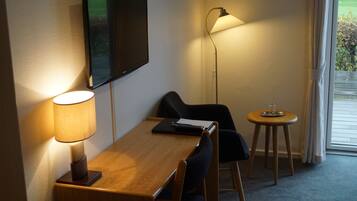 A hotel room with a wooden desk, a chair, a television mounted on the wall, a floor lamp, and a small round table with a glass on it.