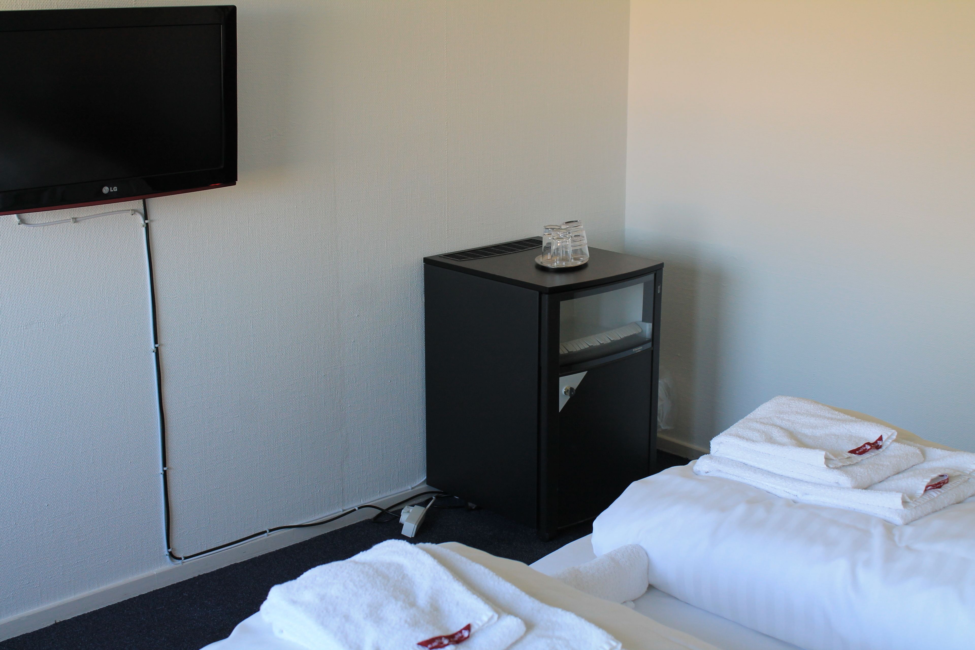 A hotel room with a flat-screen TV mounted on the wall, a small black side table with a glass top, and neatly folded white towels on the bed.