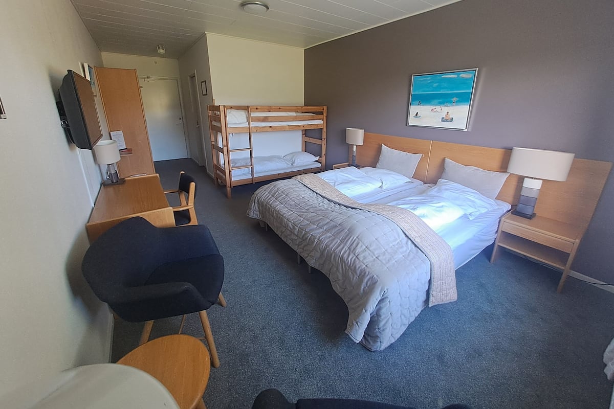 A hotel room with bunk beds, a desk, a chair, a bed with a headboard, a nightstand with a lamp, and a wall-mounted TV.