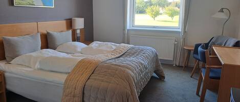 A neatly made bed with a headboard, a small table with a lamp, and a view of a park through a window.