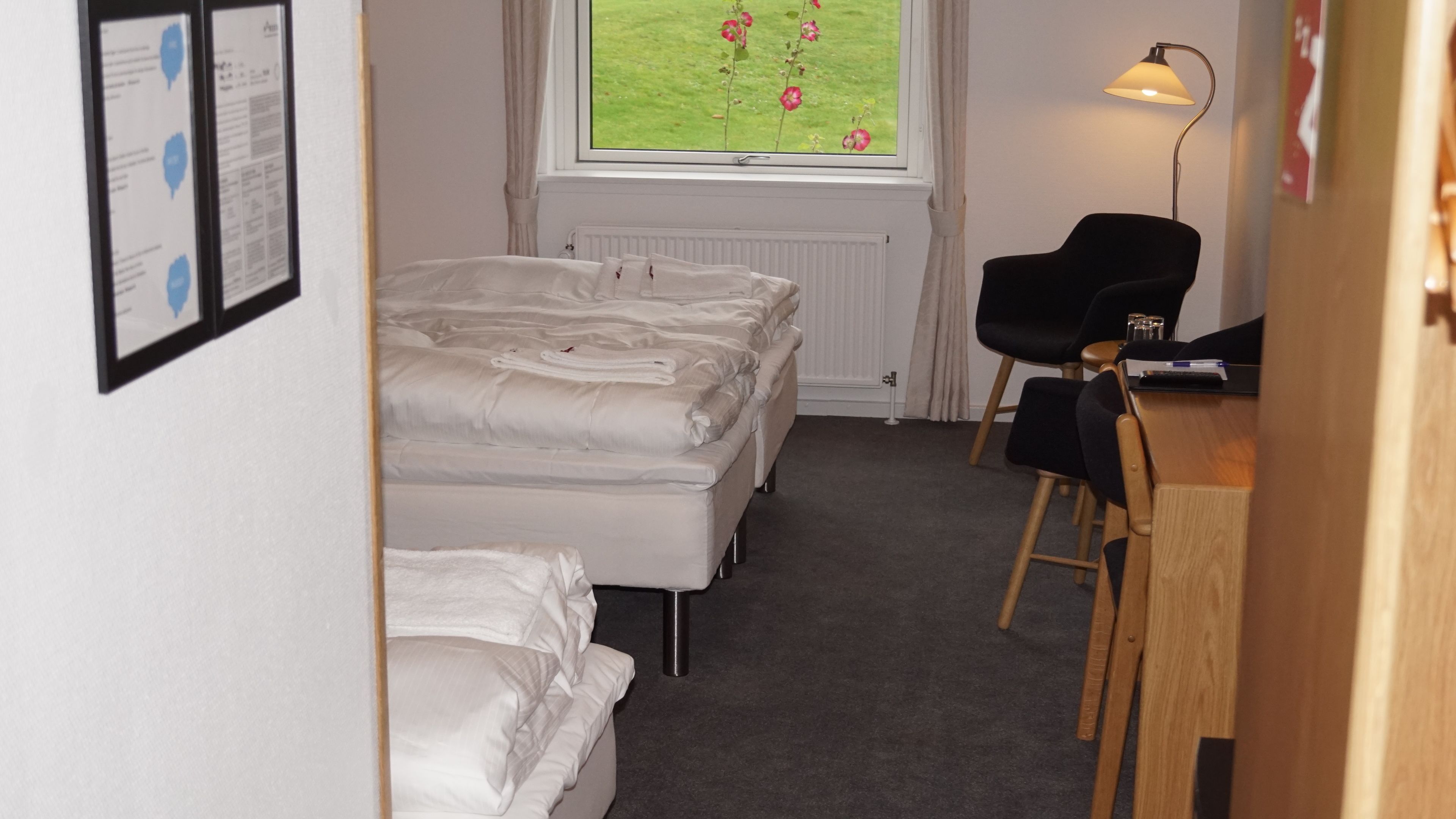 Quarto de hotel com duas camas, uma escrivaninha, uma cadeira, uma janela com vista para o verde e um radiador.