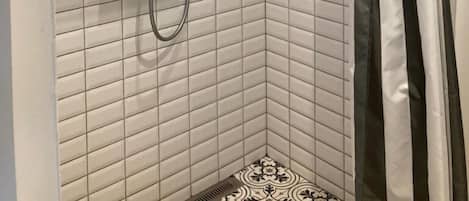 A corner shower with white tiled walls, a showerhead, a handheld shower, and a shelf with bottles.