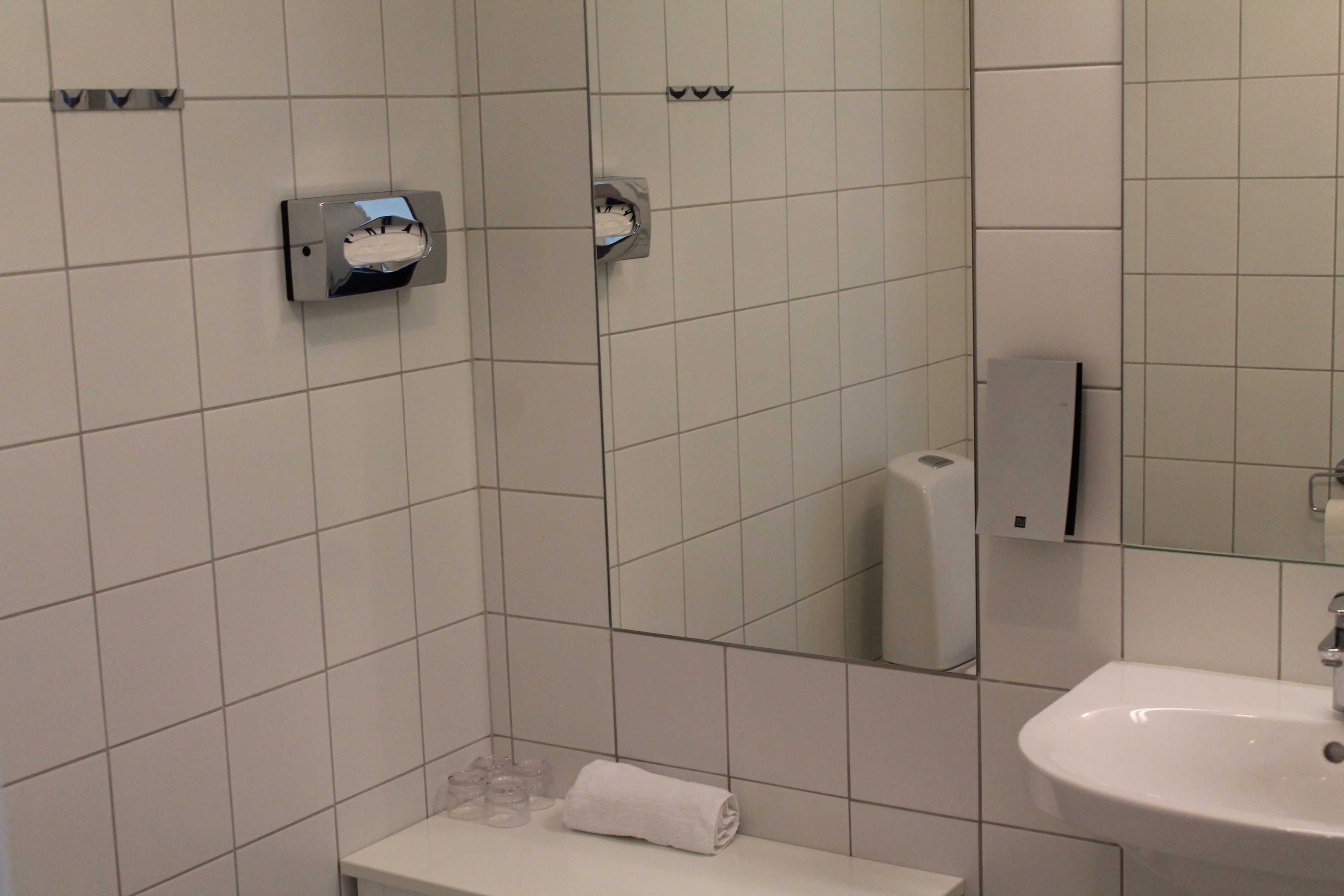 Ett badrum med vita kakelplattor, ett handfat, en spegel och en handduksdispenser.