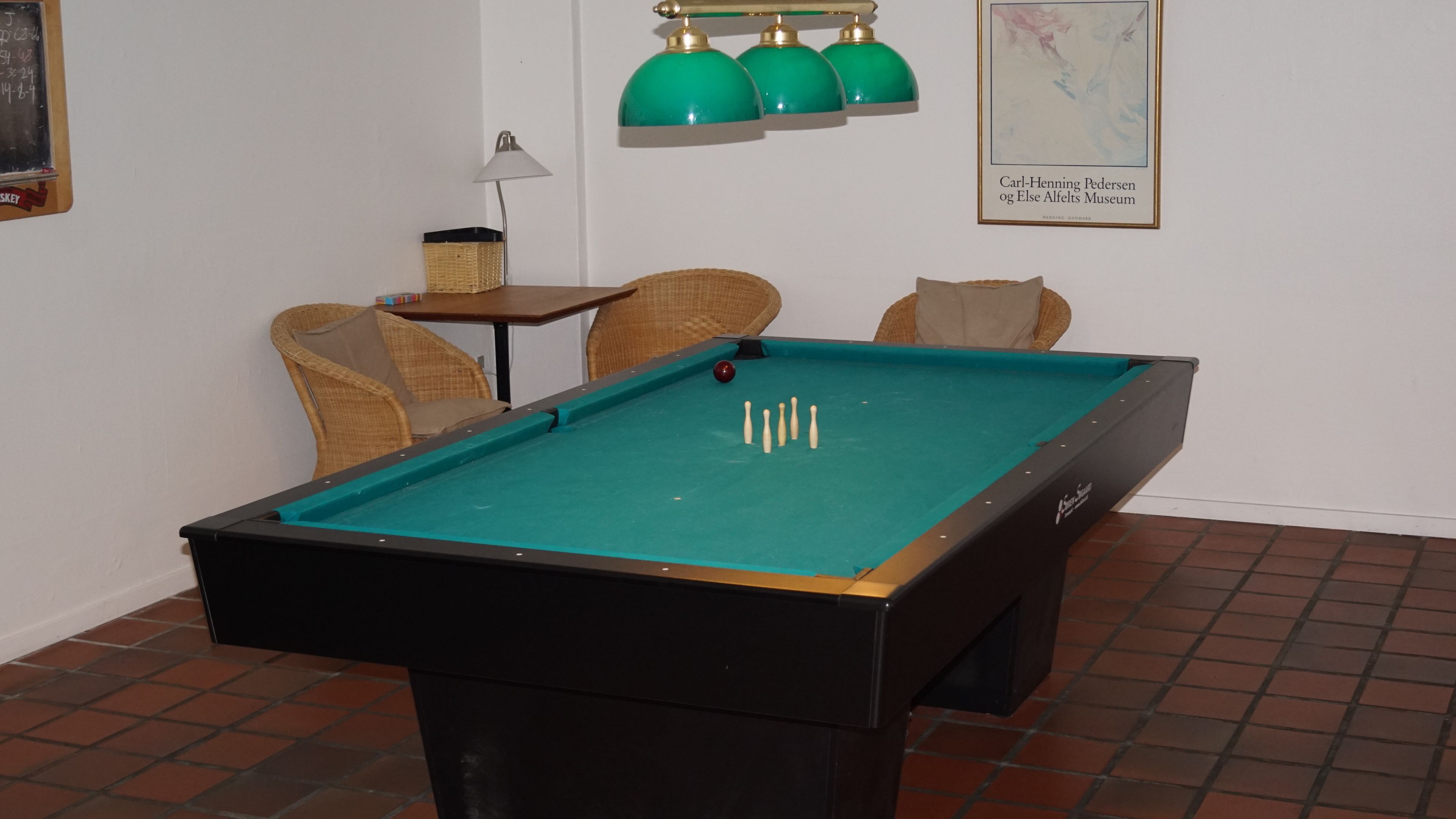 Una mesa de billar en una habitación con un mapa en la pared, una lámpara y sillas de mimbre.
