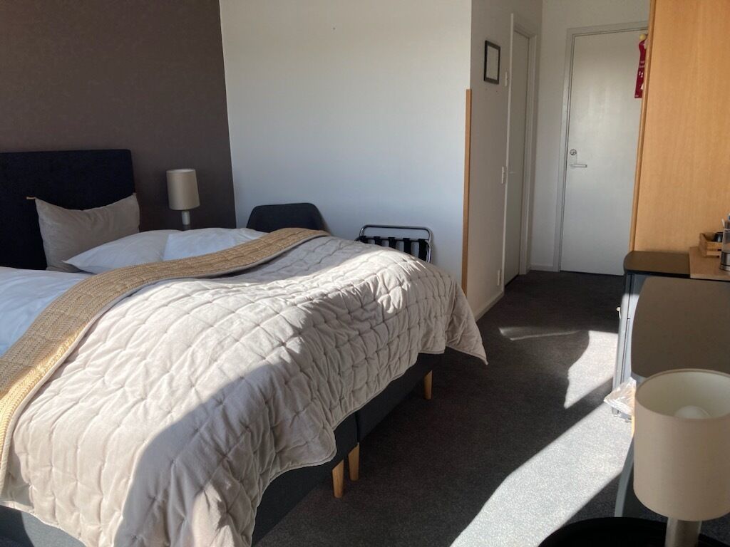 A hotel room with a bed, bedside table, lamp, and a door.