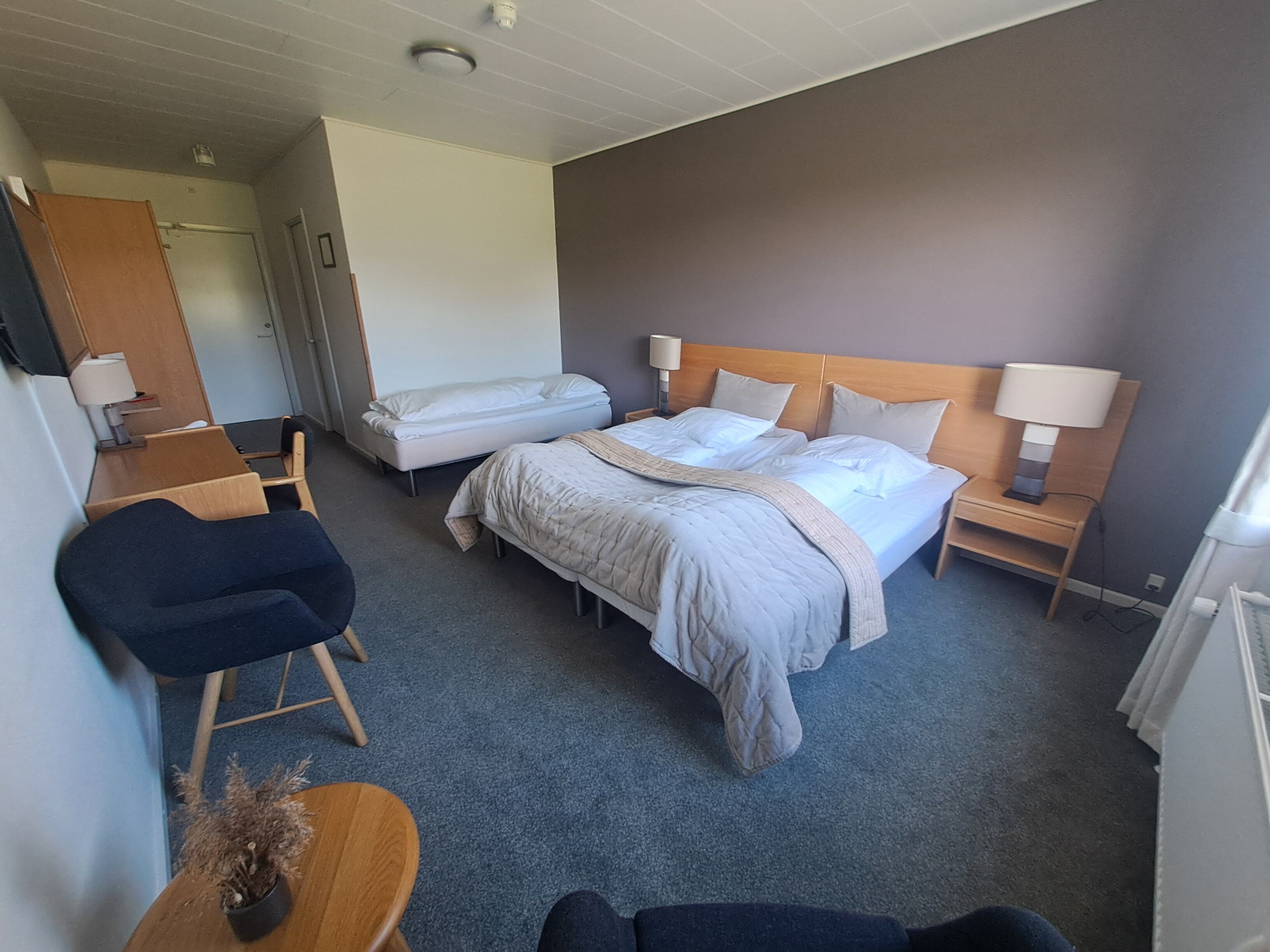 Hotellihuone, jossa on suuri sänky, kaksi yöpöytää valaisimineen, tuoli, pieni pöytä maljakolla ja ovi, joka johtaa toiseen huoneeseen.