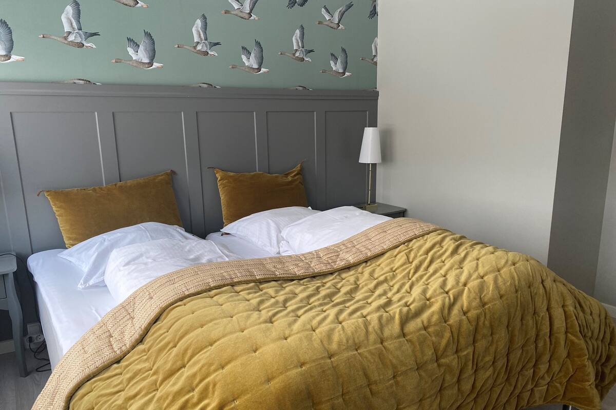 A neatly made bed with a mustard-colored quilt, two pillows, and a headboard with a patterned wallpaper of birds.