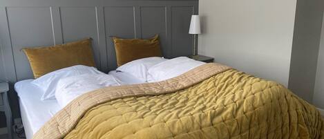 Un lit bien fait, recouvert d’une courtepointe de couleur moutarde, avec deux oreillers et une tête de lit ornée d’un papier peint à motifs d’oiseaux.