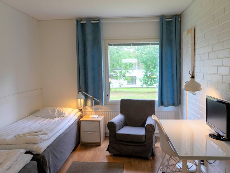 Et lille, godt oplyst værelse med en seng, et skrivebord, en stol og et fjernsyn. Der er et vindue med gardiner, en lampe og en radiator.