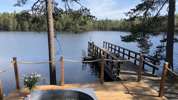 Una terraza de madera con un jacuzzi, una barandilla de cuerda y un muelle que se adentra en un lago rodeado de árboles.