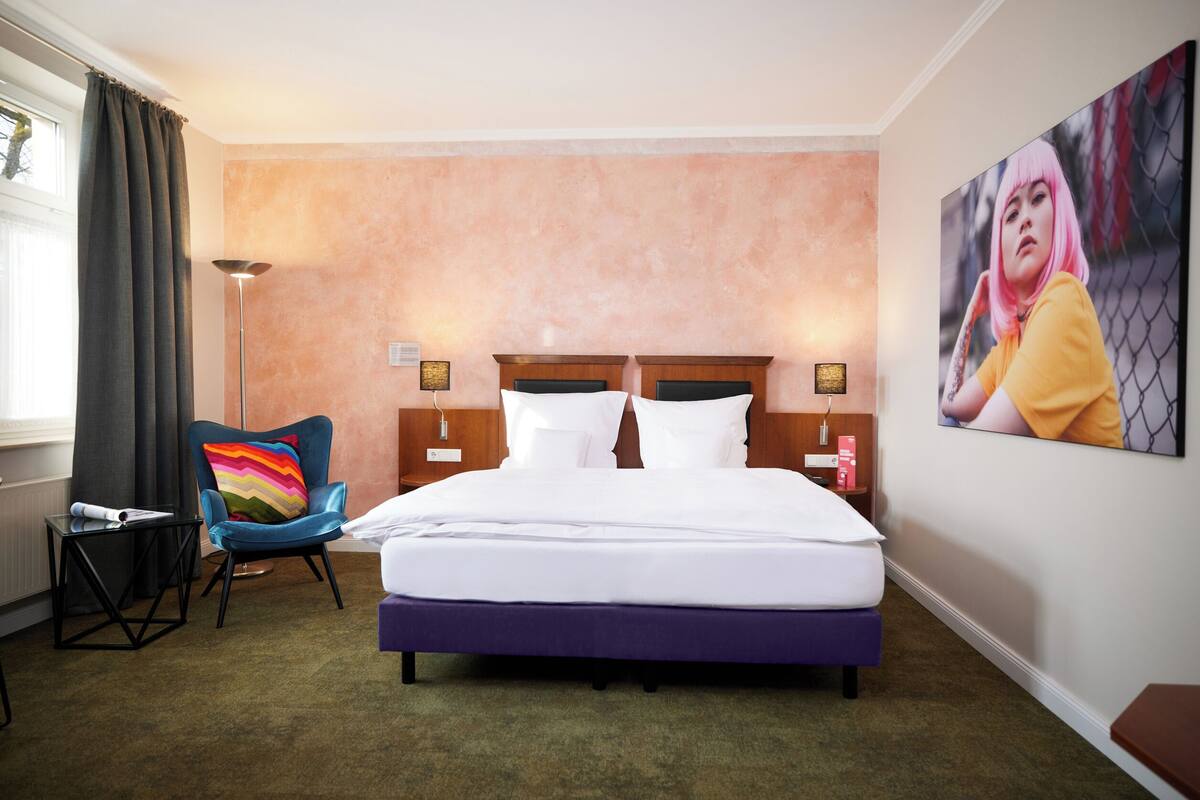 A hotel room with a large bed, a chair, a bedside table, and a painting on the wall.