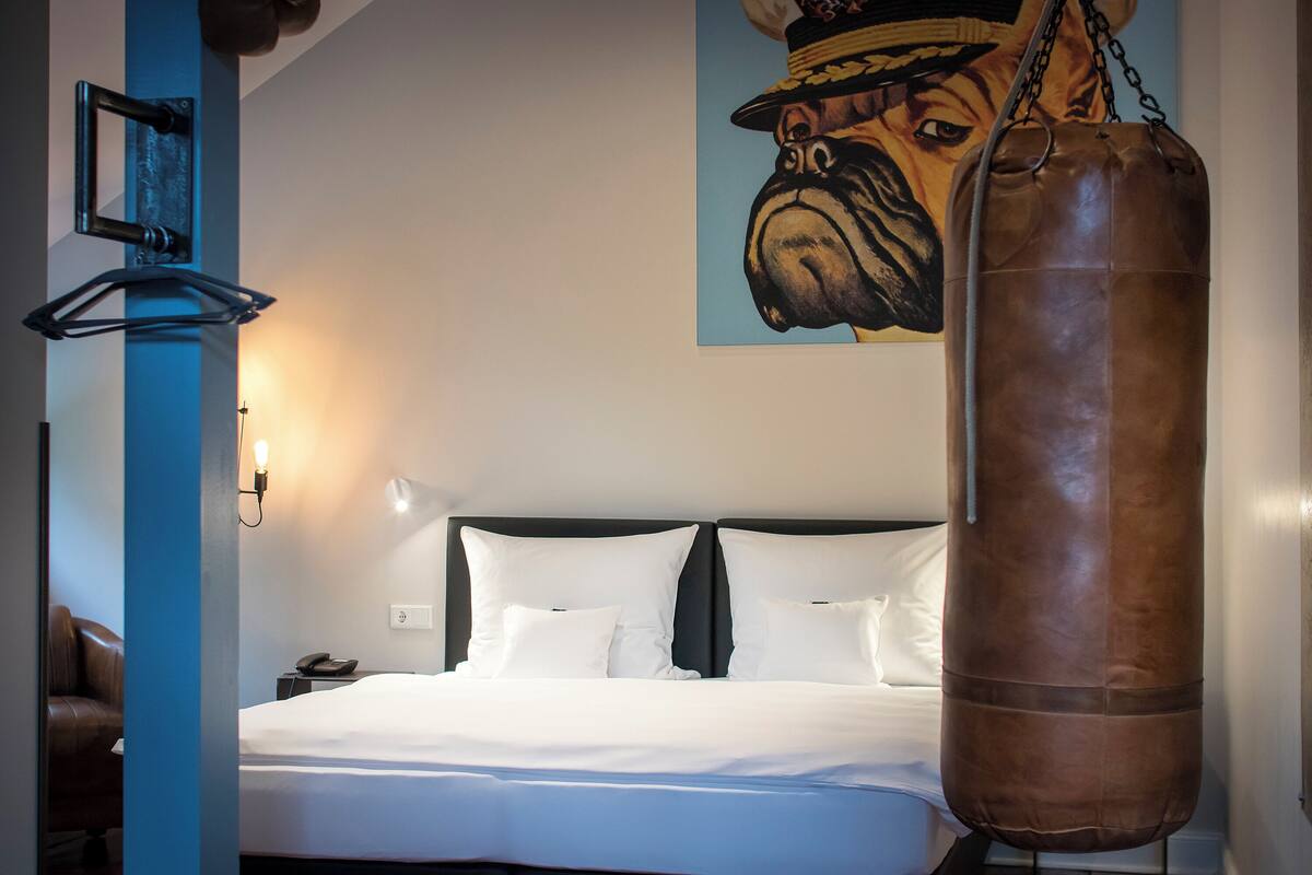 A hotel room with a bed, a punching bag, a nightstand, a telephone, and a wall-mounted light fixture.