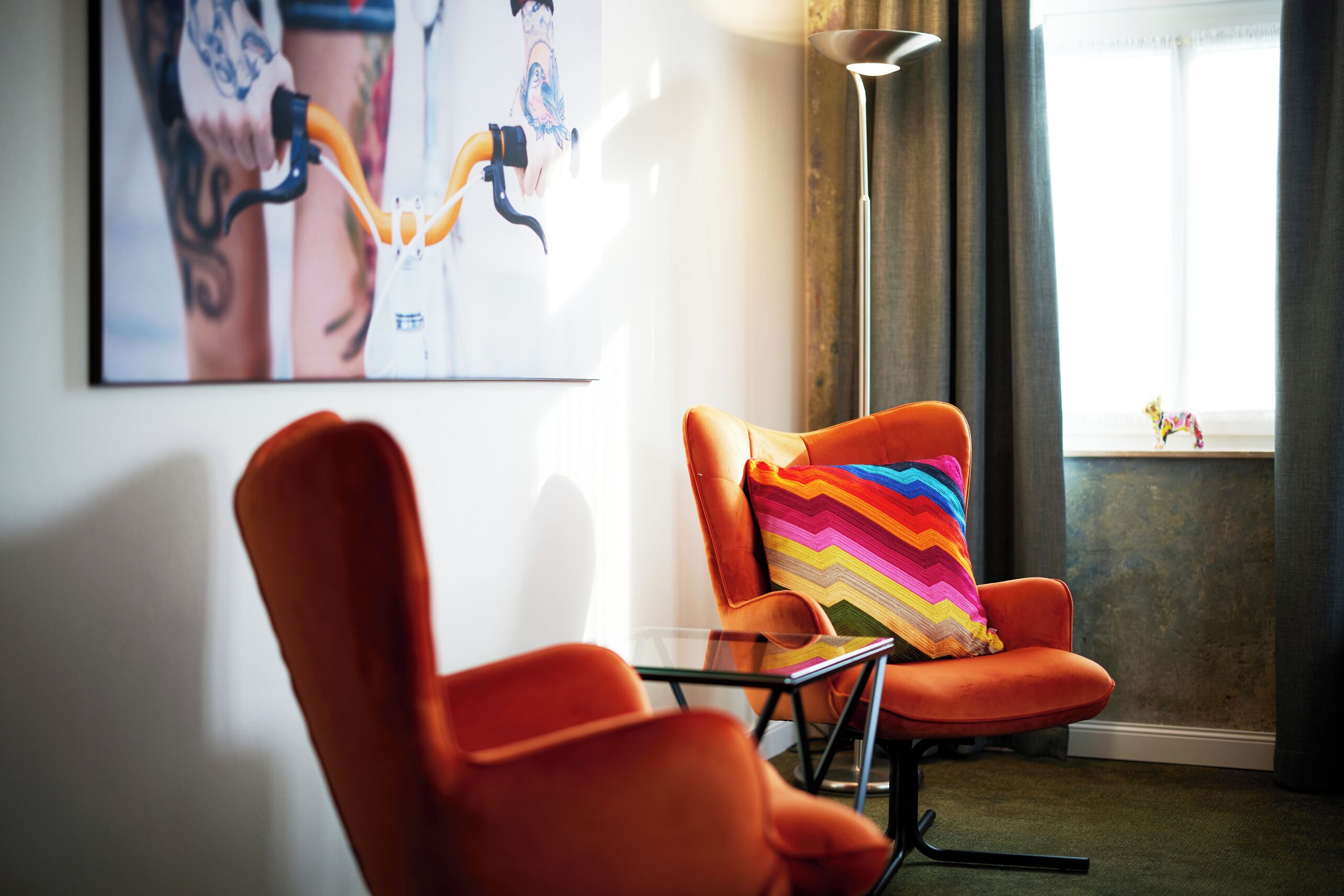 A modern living room with a glass coffee table, a vibrant striped pillow on an orange armchair, and a large framed artwork on the wall.