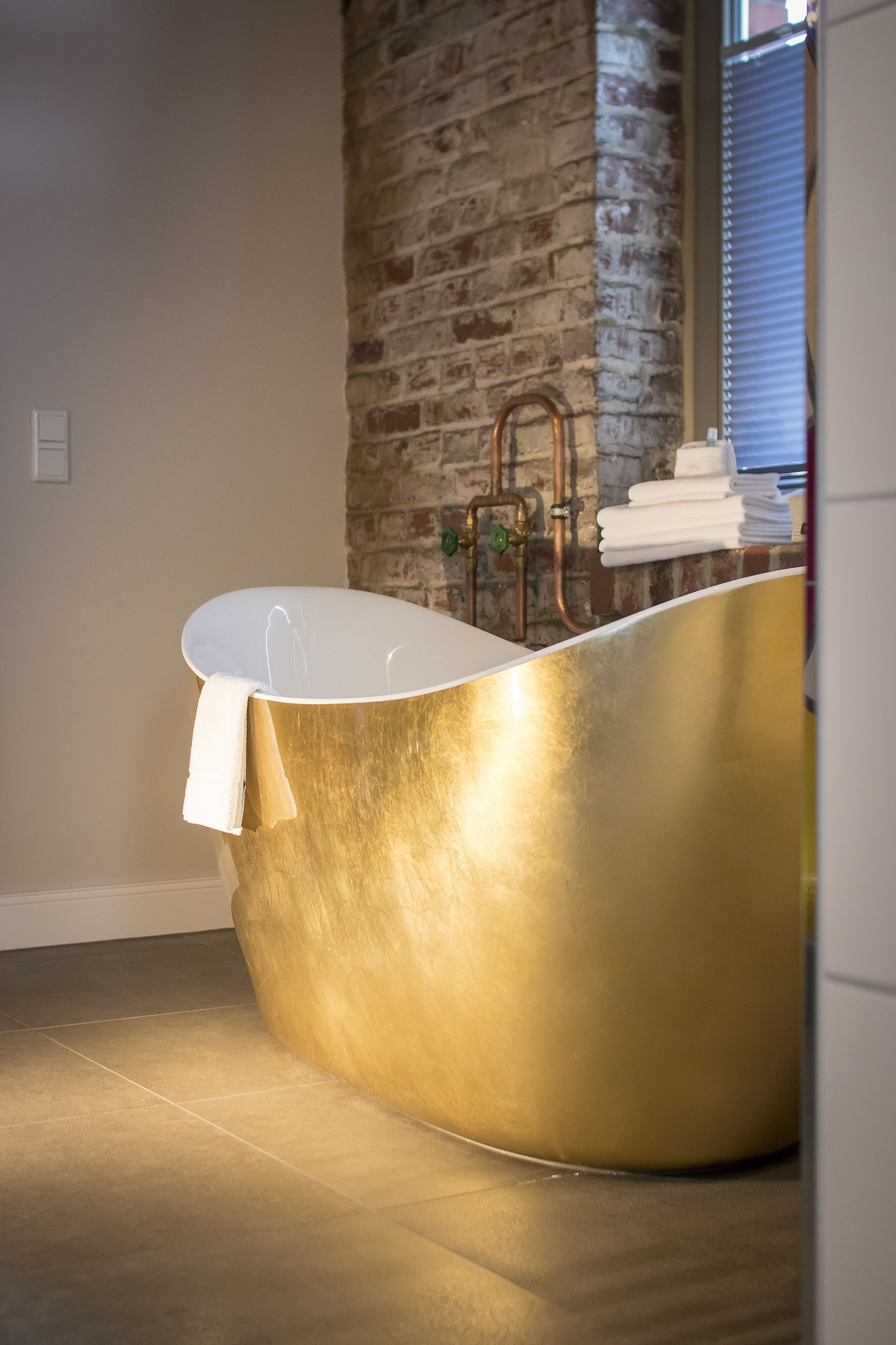 Un baño moderno con una bañera grande de color dorado independiente, una pared de ladrillo y una ventana con persianas.