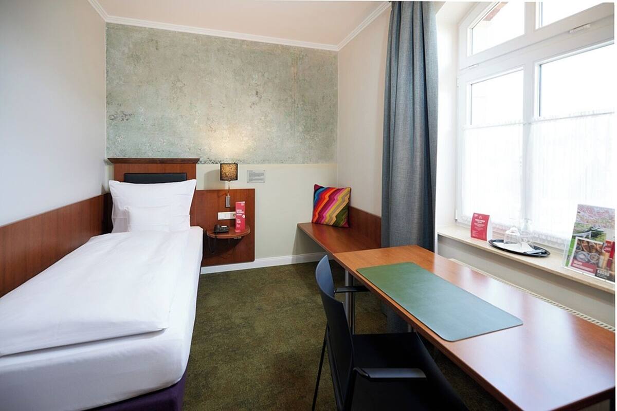 A hotel room with a bed, a desk, a chair, a small table, a nightstand, a lamp, a colorful pillow, a window with curtains, and a view of the outside.