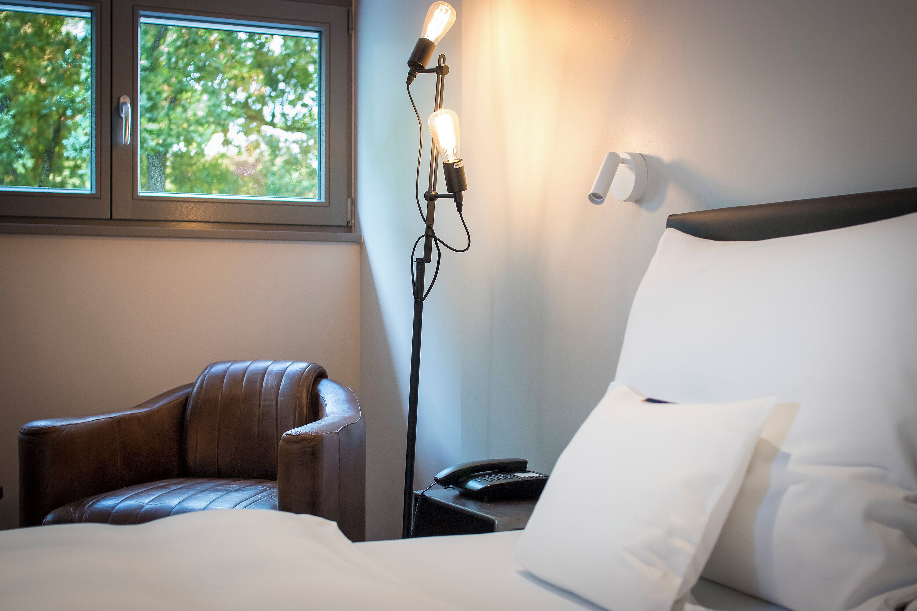 A hotel room with a bed, a leather armchair, a bedside table with a lamp, and a window with a view of trees.
