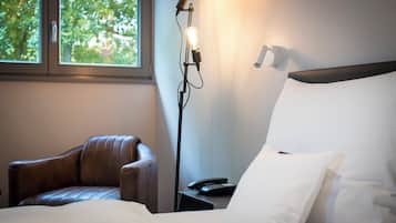 Una camera d'albergo con un letto, una poltrona in pelle, un comodino con una lampada e una finestra vista alberi.