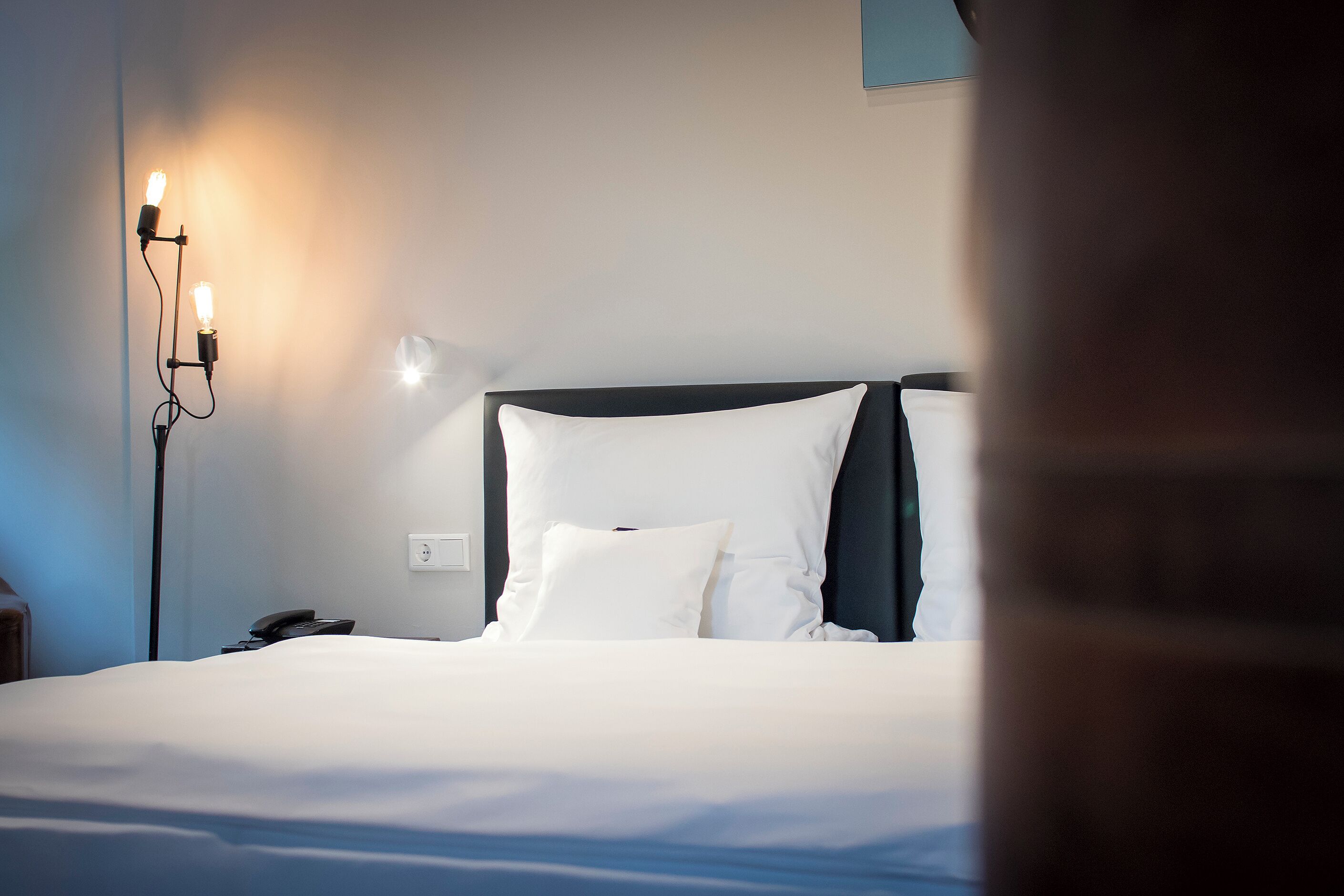 A neatly made bed with white linens, a bedside telephone, and a wall-mounted light fixture.