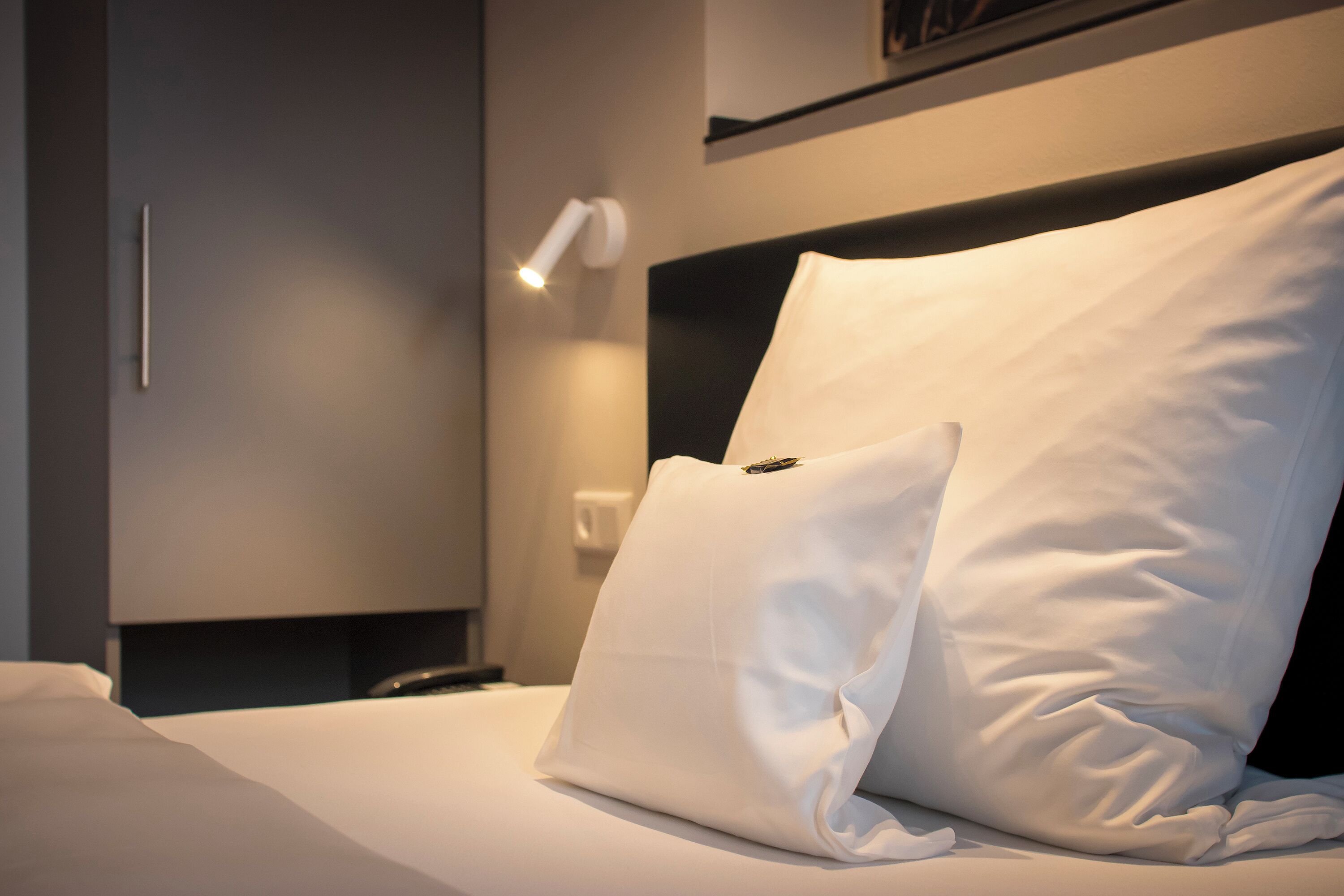 A neatly made bed with white linens and pillows, a bedside lamp, and a modern headboard.