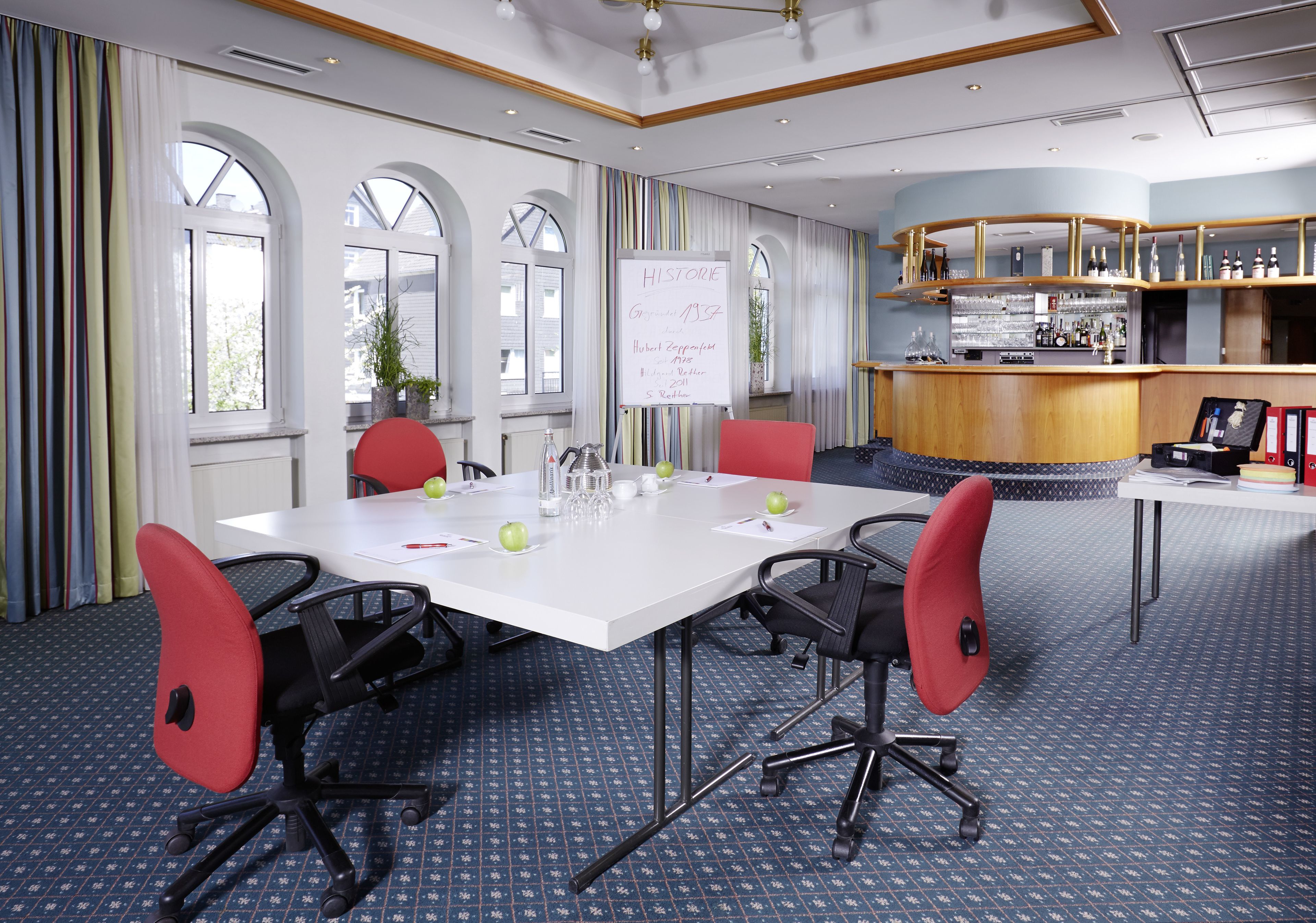 A conference room with a white table, red chairs, a whiteboard, and a bar area with shelves of bottles.