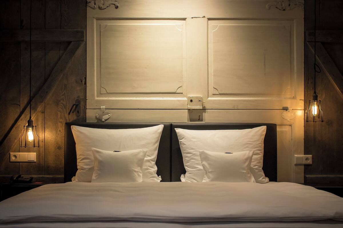 A neatly made bed with white linens and pillows, a wooden headboard, and wall-mounted light fixtures.