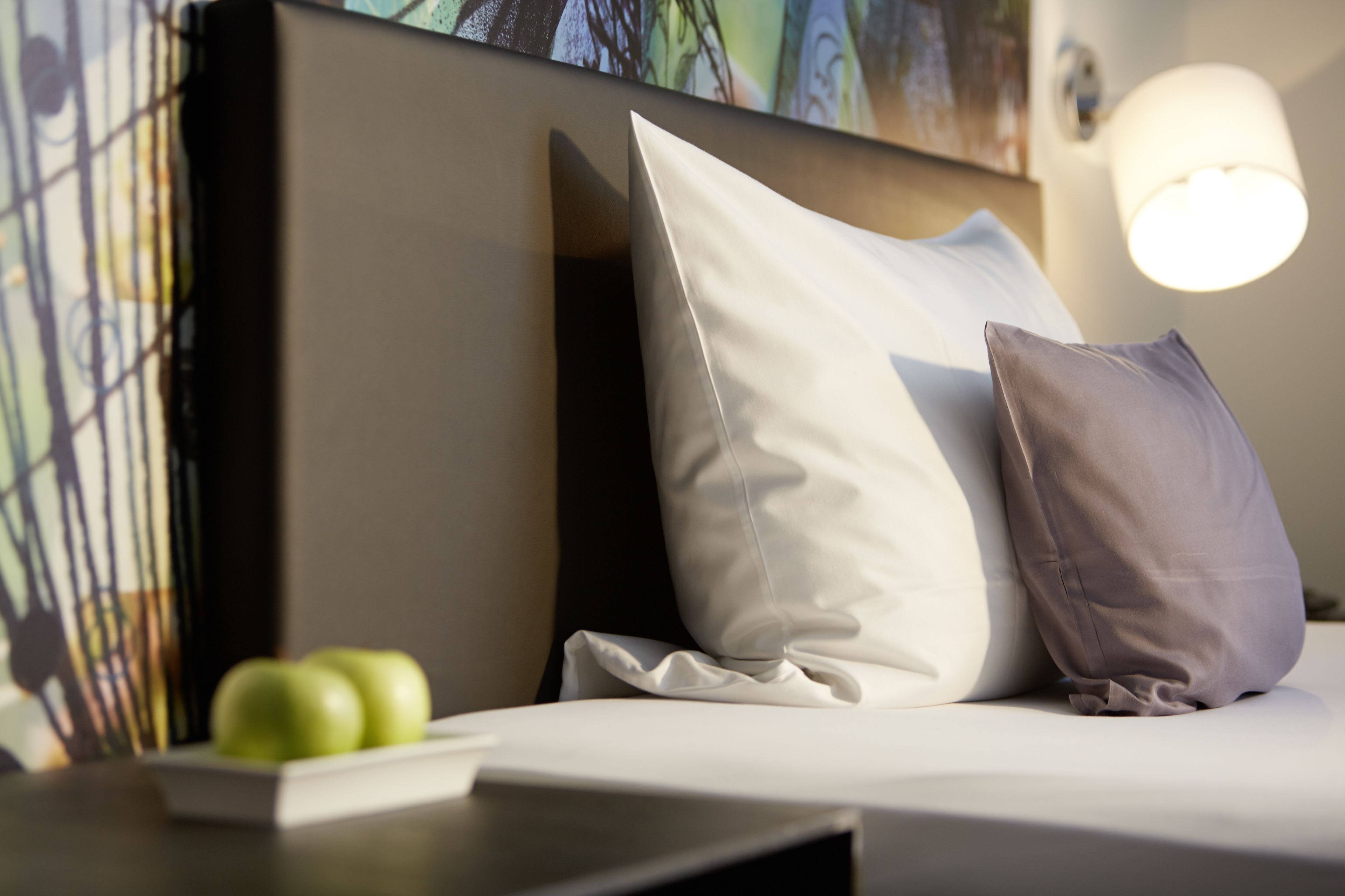 Una cama bien tendida con almohadas blancas y grises, una mesita de noche con un libro y dos manzanas, y una lámpara fijada a la pared.