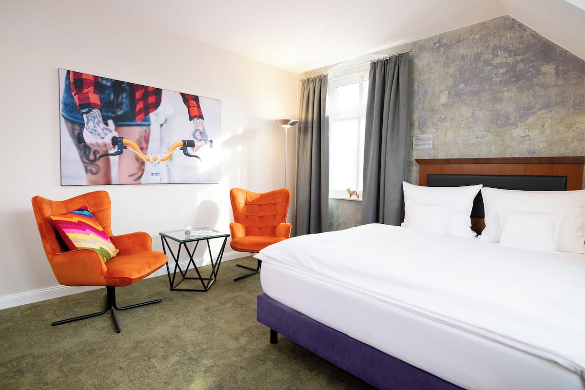 A hotel room with a large bed, two orange armchairs, a small table, and a painting on the wall.