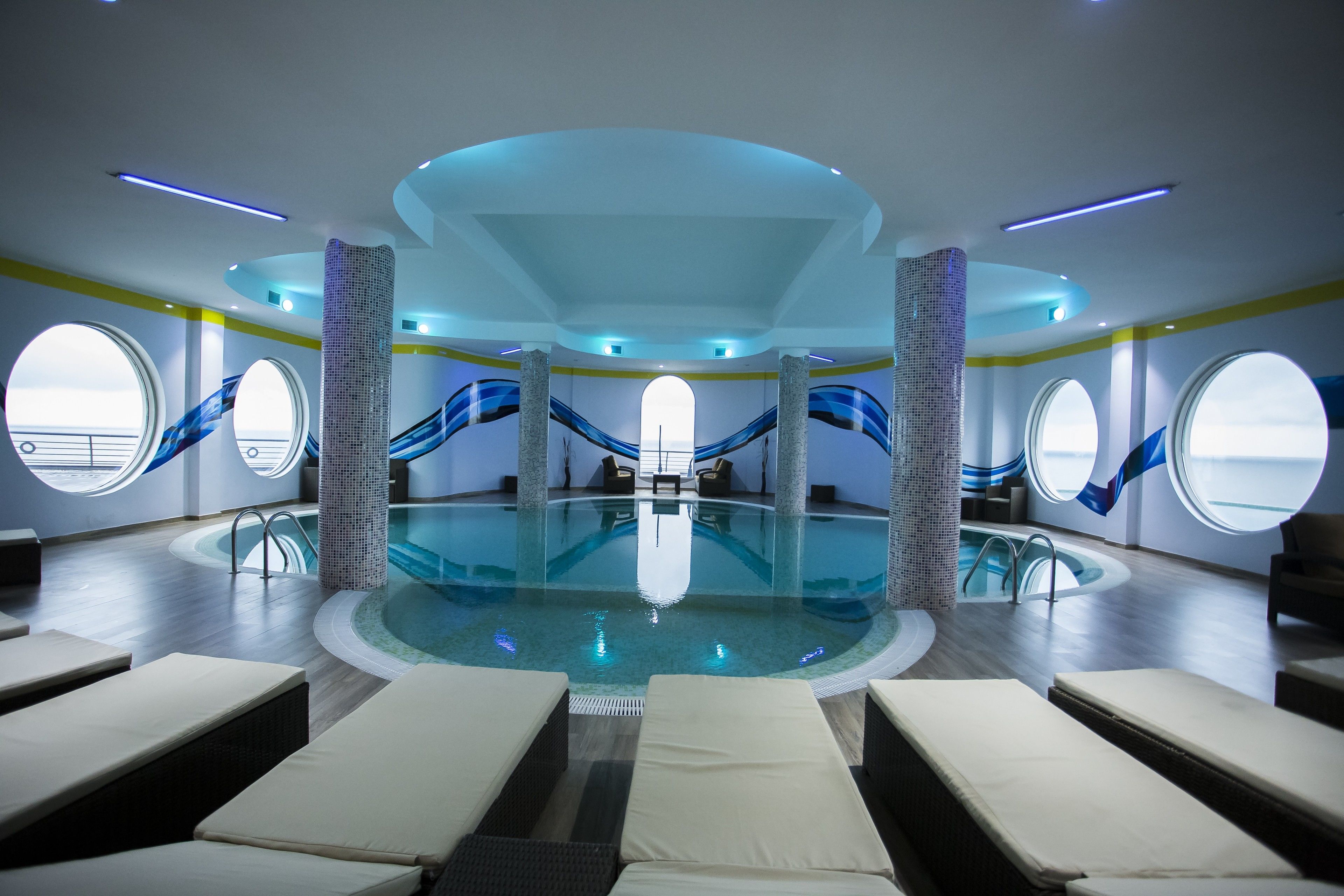 A modern indoor pool area with a central pool, lounge chairs, and a view through large windows.