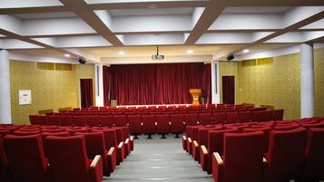 Un teatro con hileras de asientos rojos, un escenario con cortina roja y un atril.