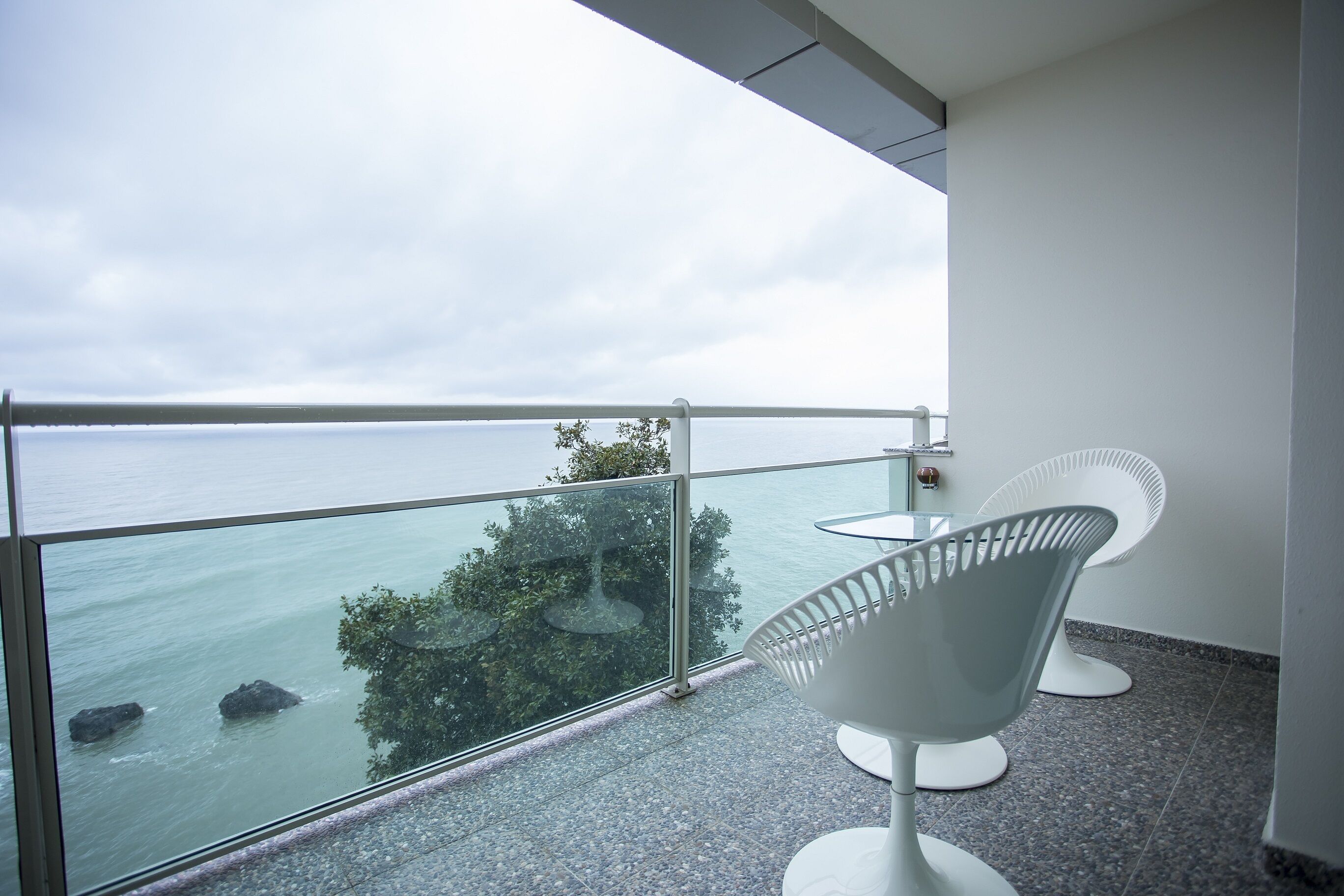 A balcony with a glass railing, two white chairs, and a view of the sea and trees.