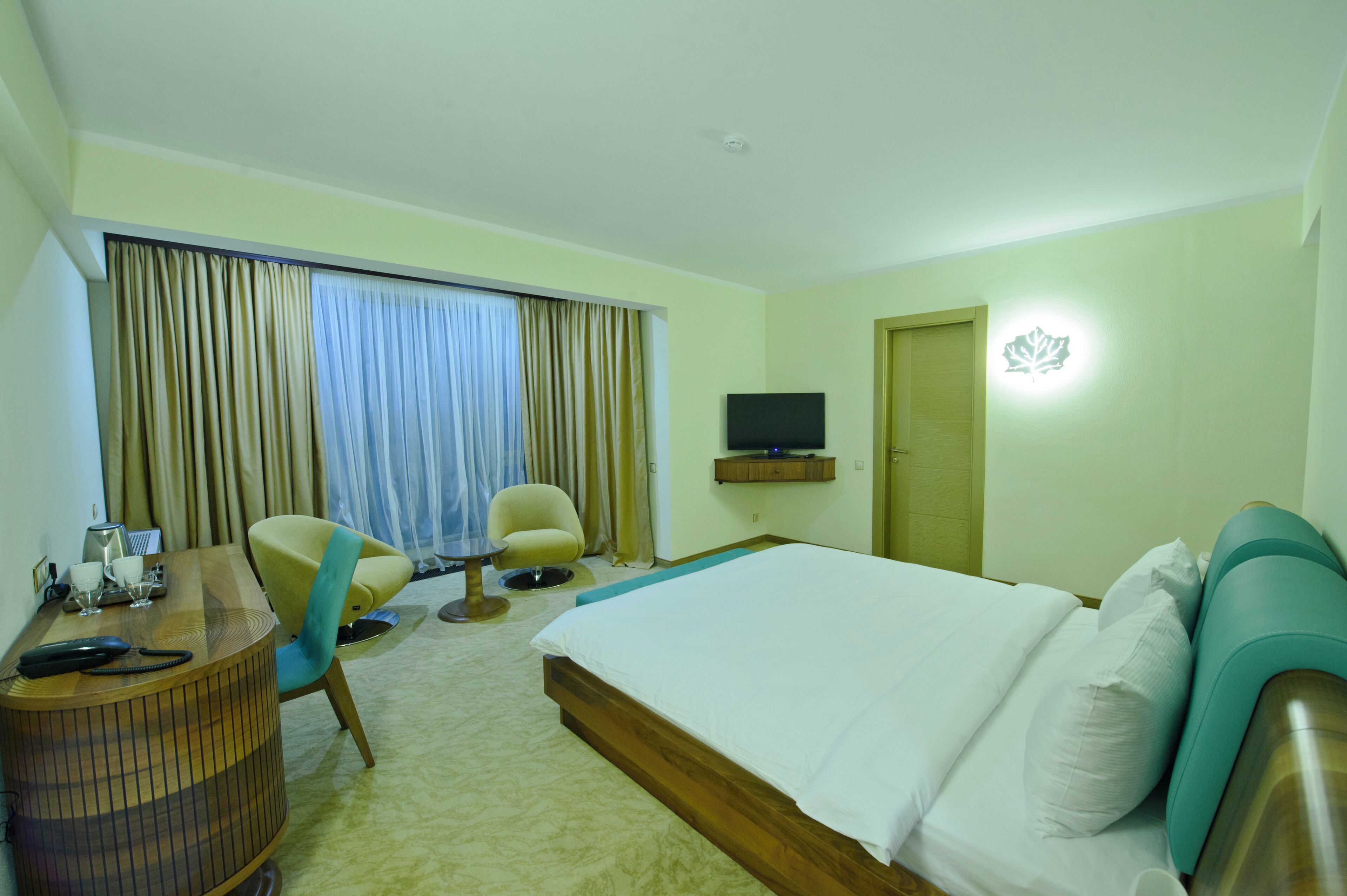 Quarto de hotel com cama, escrivaninha, cadeira, televisão e janela com cortinas.