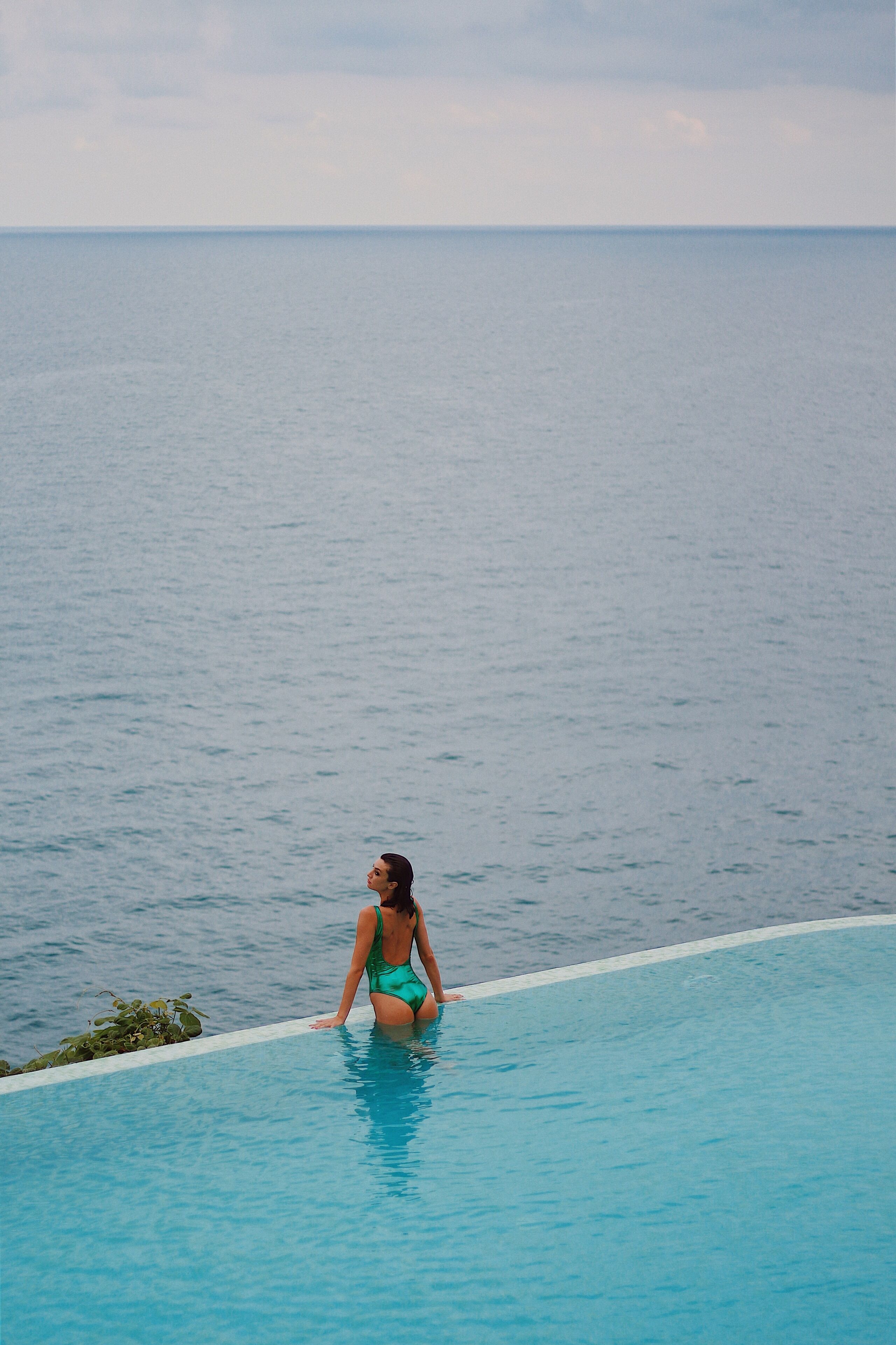 A person in a blue swimsuit at the edge of a pool overlooking the sea.