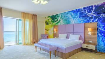 A modern hotel room with a large bed, a bench, a view of the sea, and a colorful abstract wall.