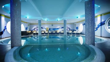 Una piscina interior con paredes de azulejos y columnas, sillas de descanso y una zona de estar circular.