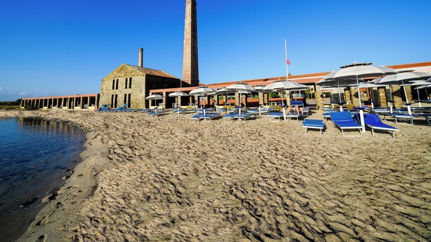 Une plage bordée d’une rangée de chaises longues et de parasols, un bâtiment en pierre et une haute cheminée.