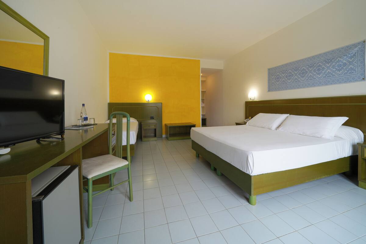 Een hotelkamer met een bed, een bureau met televisie, een stoel, een nachtkastje en een wandlamp.