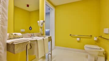 Un baño con paredes amarillas, un lavamanos blanco, un espejo, un perchero para toallas, un secamanos, un inodoro y una barra de apoyo.
