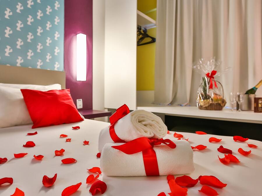 Cama bem arrumada com pétalas de rosa vermelhas espalhadas sobre o edredom, uma toalha branca enrolada com fita vermelha e uma pequena cesta de presentes com fita vermelha em uma mesa de cabeceira.