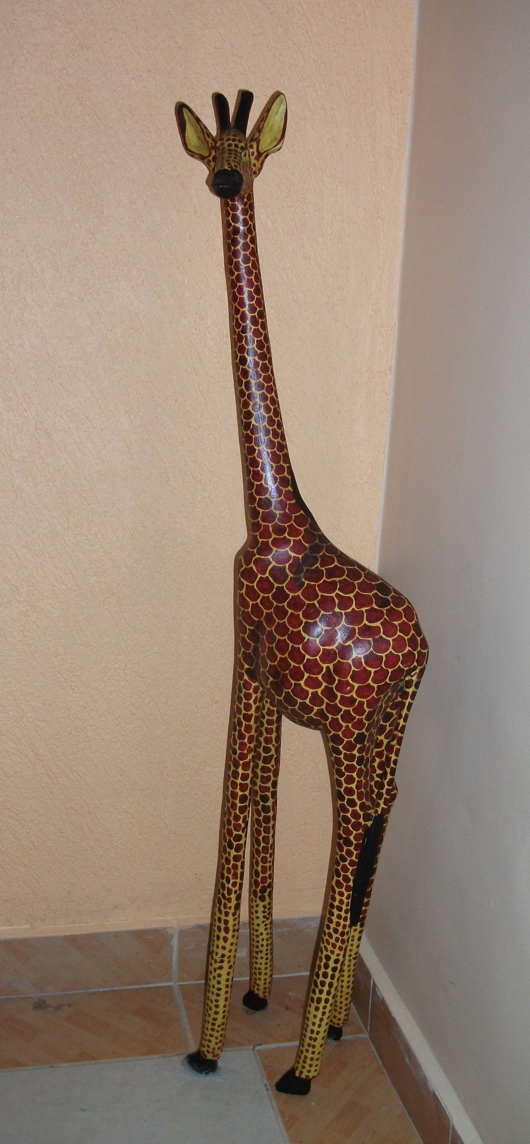 A giraffe statue in a corner of a room with beige walls and tiled flooring.