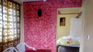 A room with a red patterned wall, a bed with a canopy, a small wooden table, and a white plastic chair with a purple cushion.