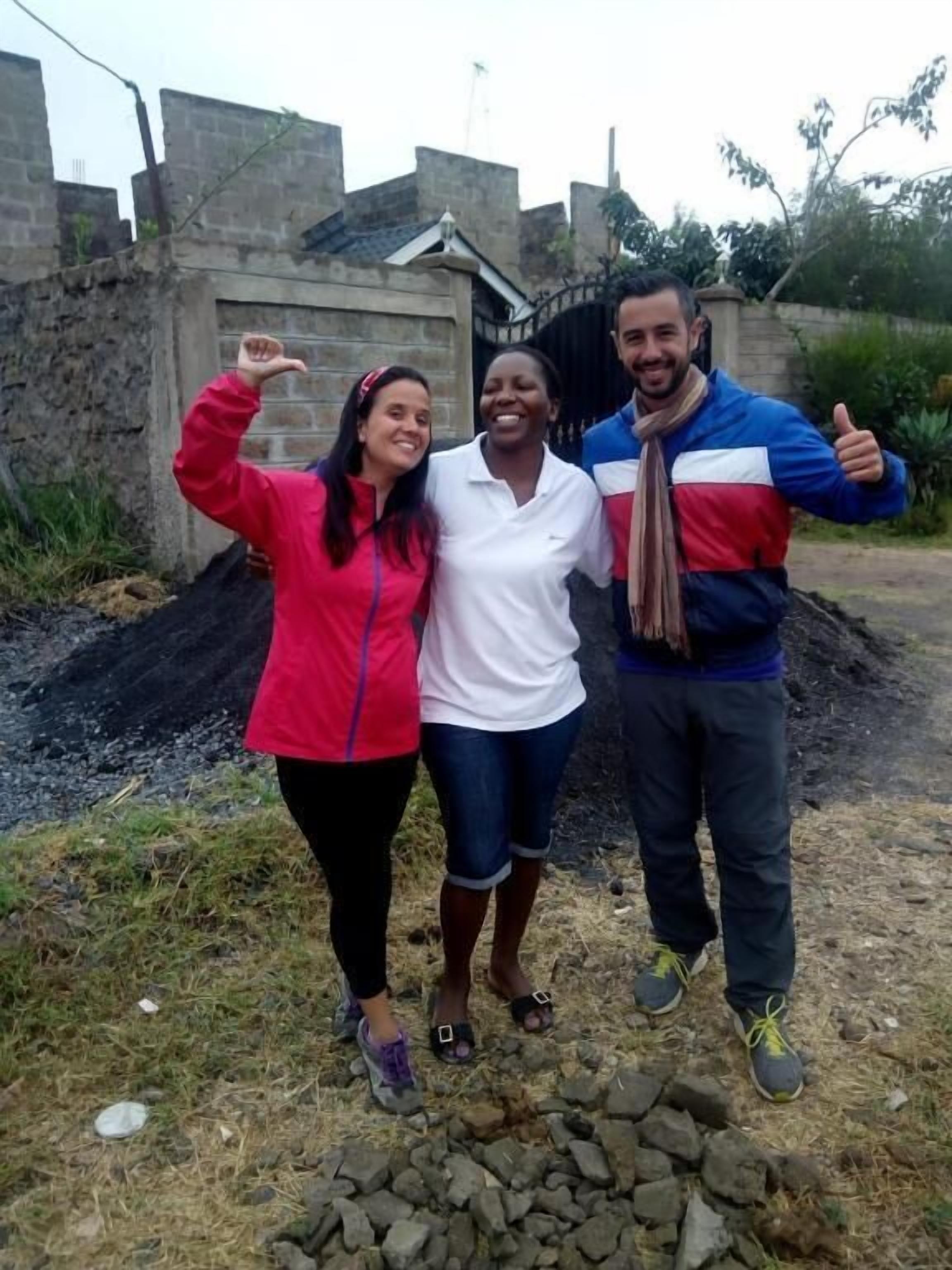 Tres personas de pie al aire libre, con un muro de piedra y un edificio al fondo.
