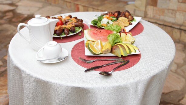 A round table with a white tablecloth, a white teapot, and a plate of food including watermelon, pineapple, and other fruits. - Sinai Suites Hotel (Kigali)