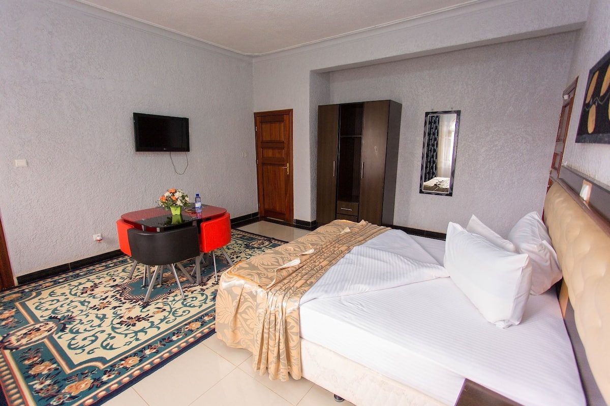 A hotel room with a bed, a TV mounted on the wall, a desk with a chair, and a wardrobe.