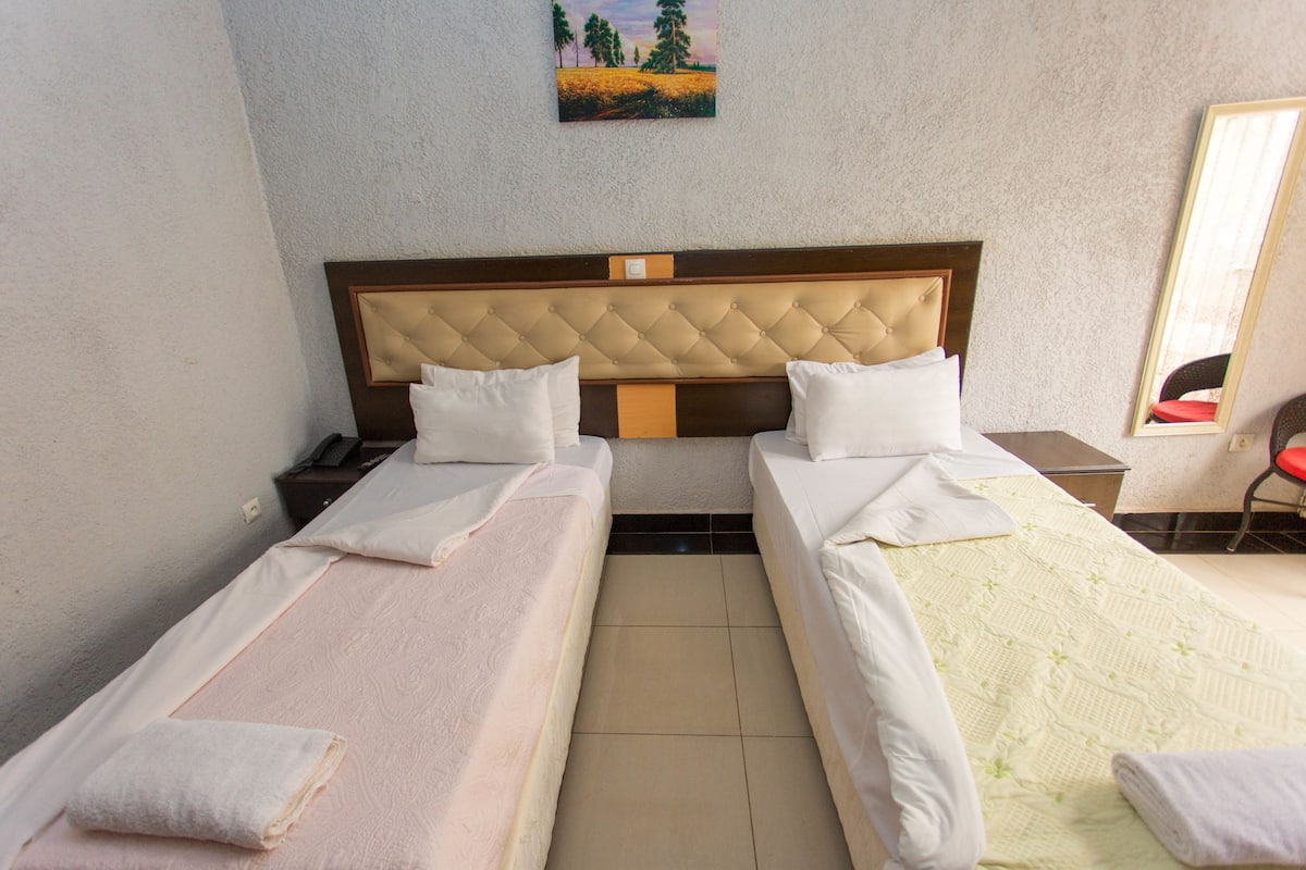 A hotel room with two single beds, a wooden headboard, a nightstand, a mirror, and a painting on the wall.