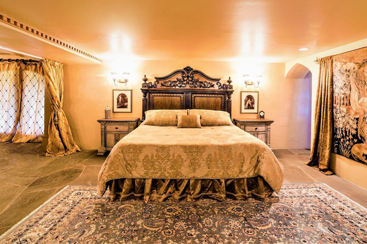 A large bed with a wooden headboard, two bedside tables with lamps, a patterned rug, and a tapestry on the wall.