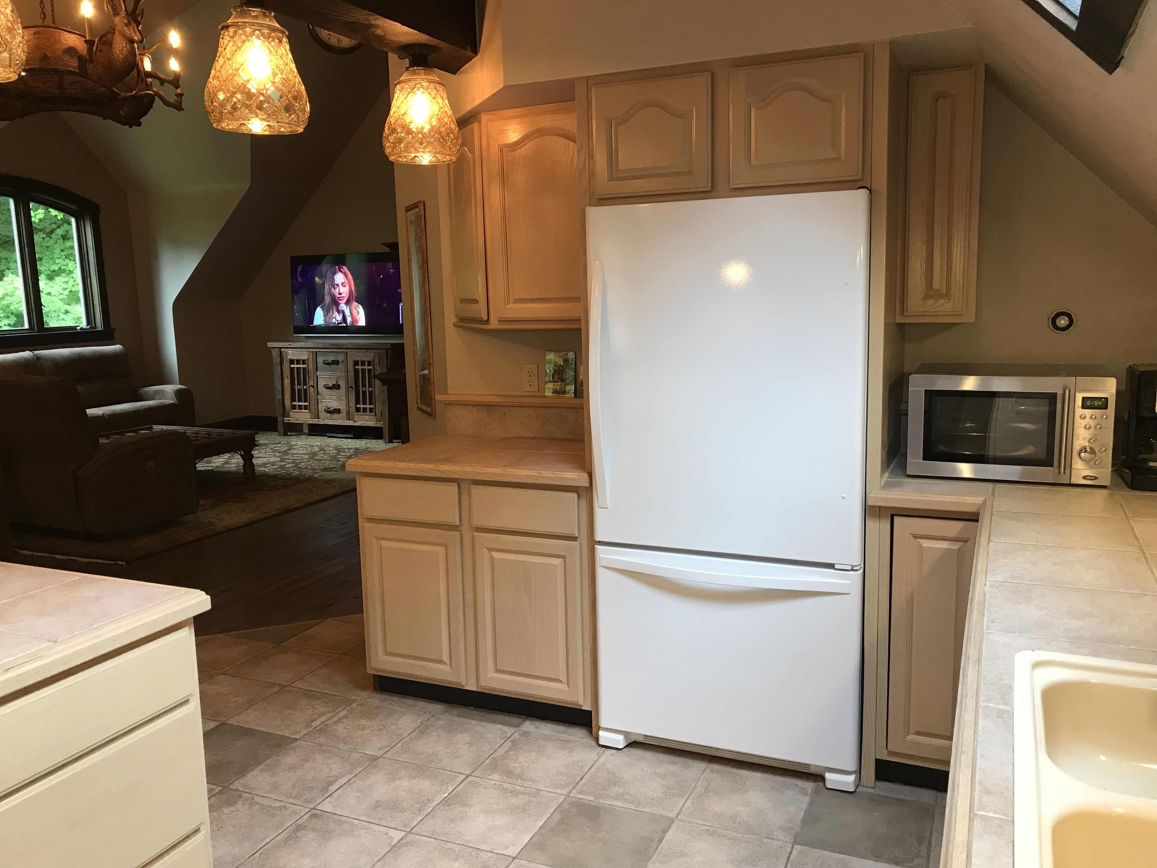A kitchen with a white refrigerator, a microwave, and wooden cabinets. There is a dining area with a couch and a TV in the background.
